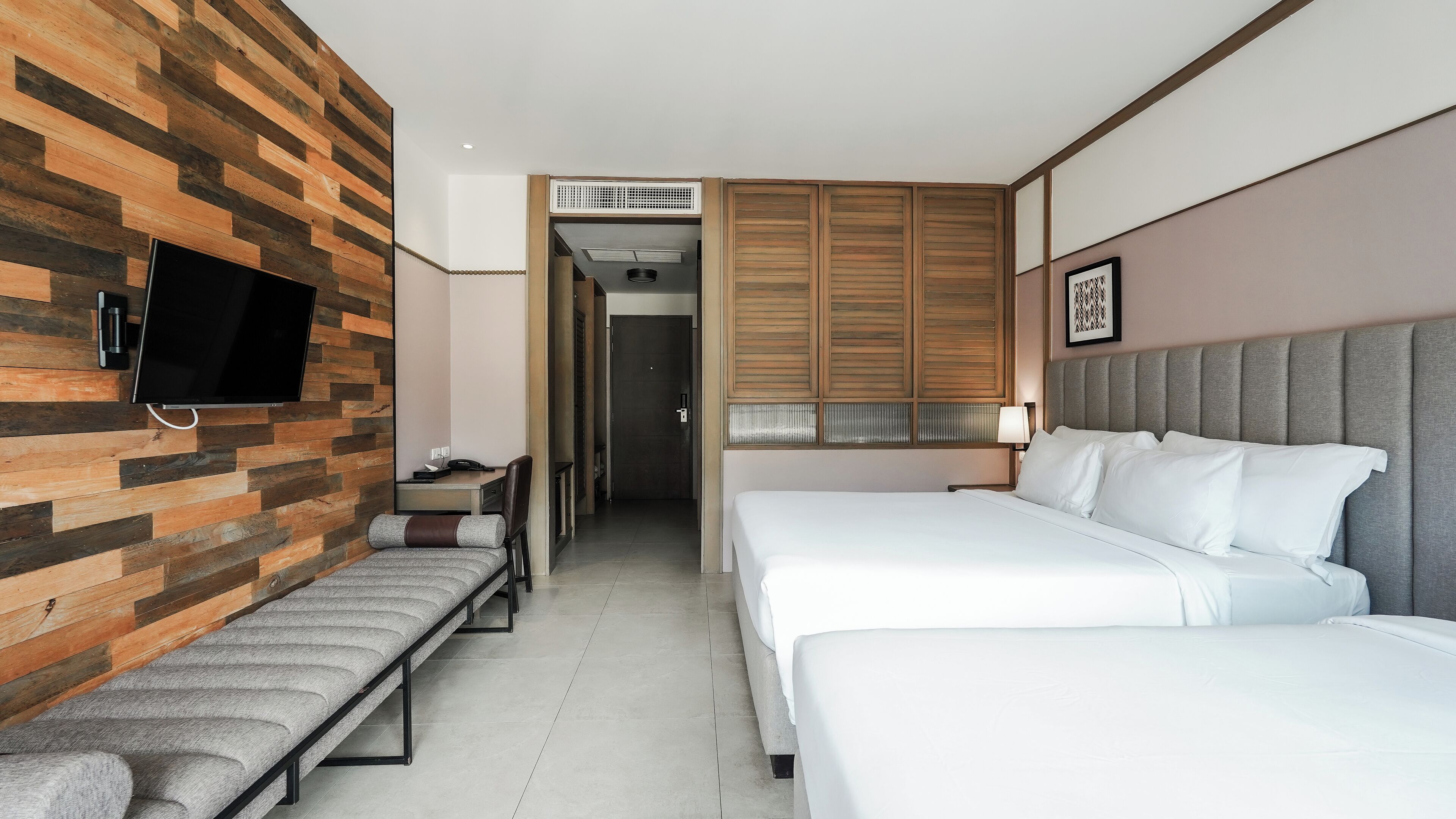 A hotel room with two beds, a bench, a TV mounted on a wooden wall, and a door leading to a bathroom.