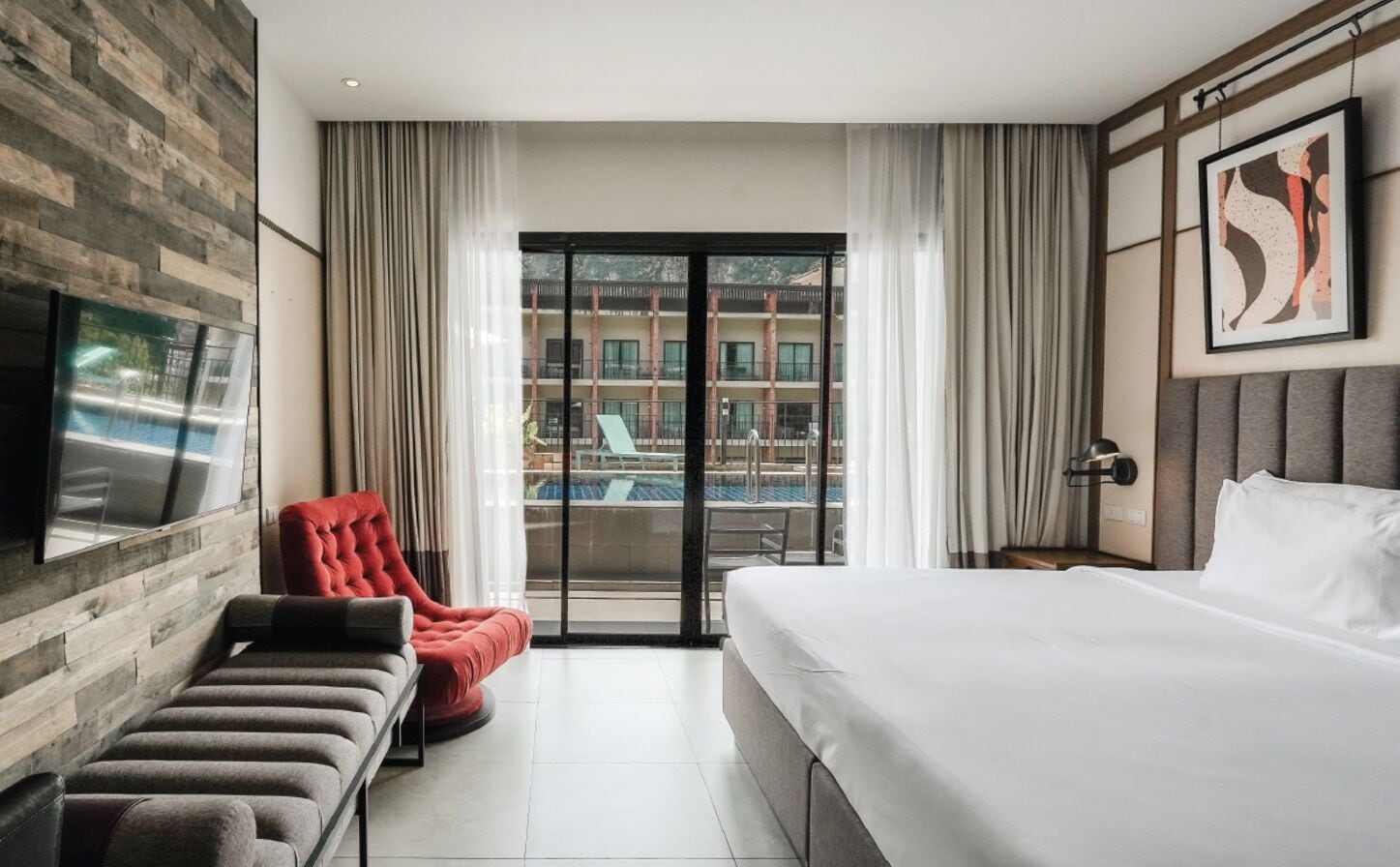 Hotelkamer met een bed, een bankje, een rode stoel, een televisie en uitzicht op een zwembad en gebouwen.