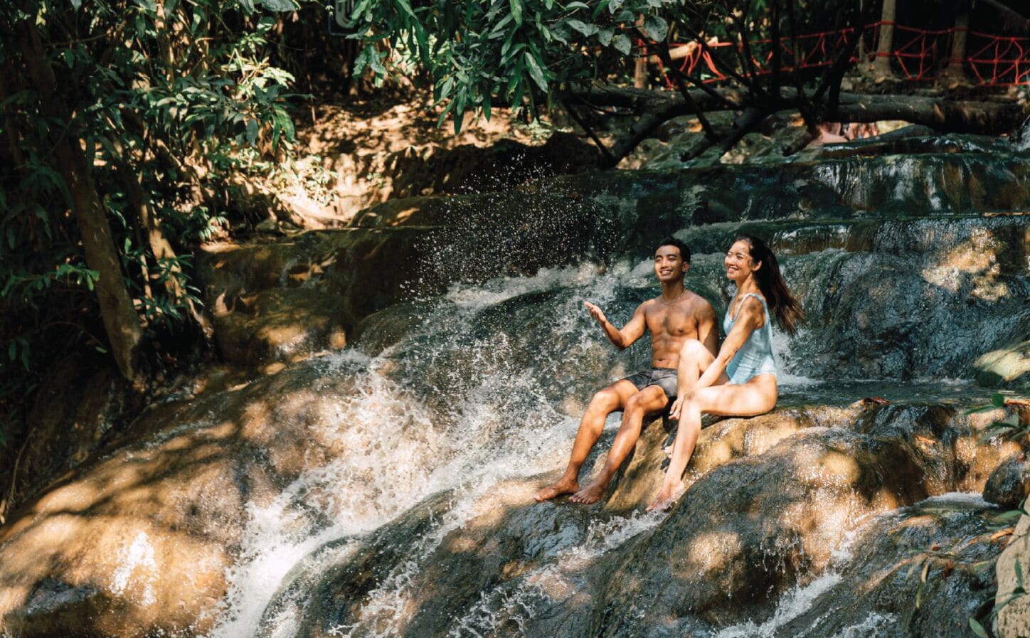 Dos personas sentadas sobre un saliente rocoso, con una cascada al fondo.