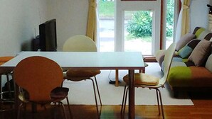 A dining area with a table and chairs, a sofa, and a window with curtains. - Guesthouse Annabel (Hakodate)