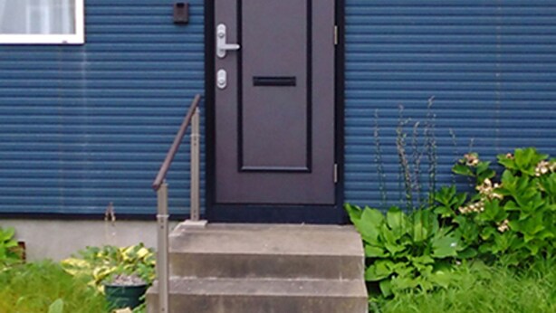 A blue building with a dark door, a small window with a curtain, and a security camera. - Guesthouse Annabel (Hakodate)