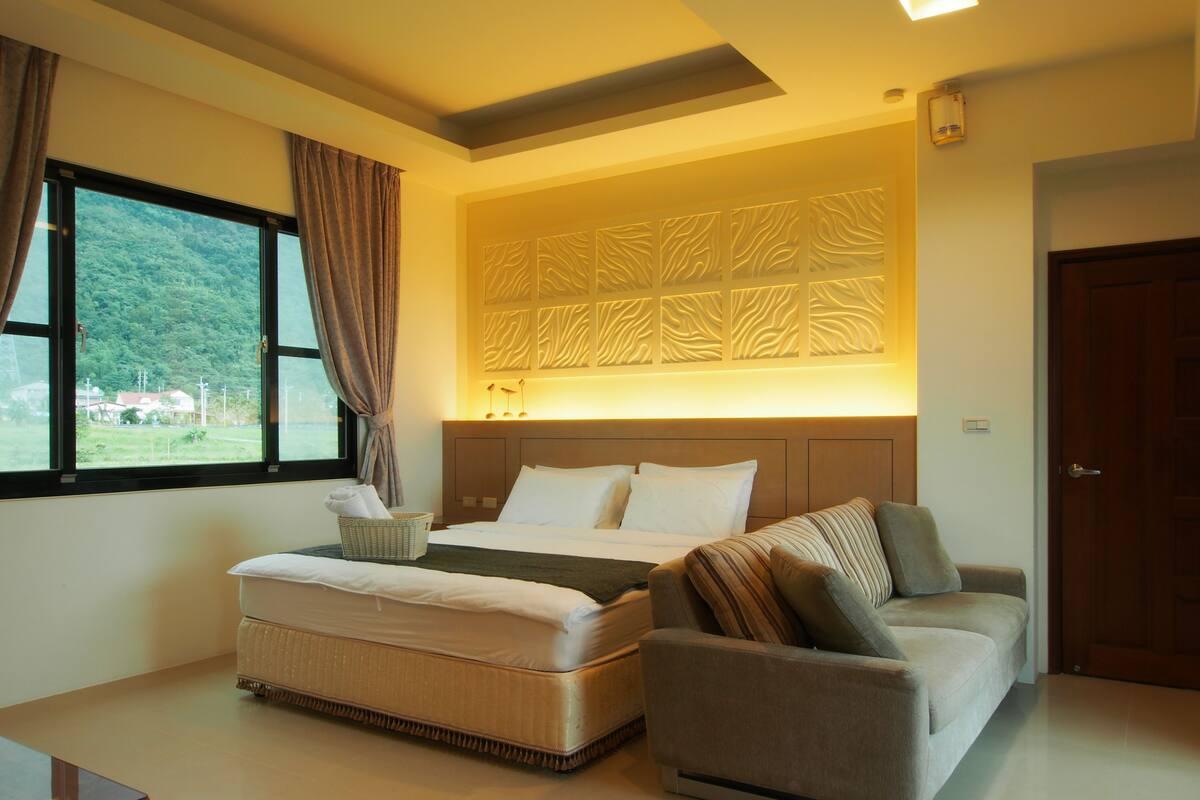 A bedroom with a bed, a sofa, a window with curtains, a wall with a decorative panel, and a door.