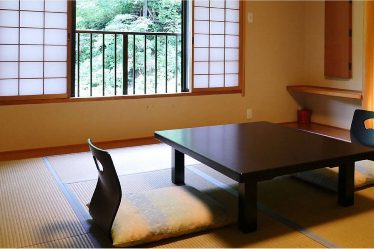 A traditional Japanese room with tatami flooring, a low table, a chair, and a view of greenery through a window.