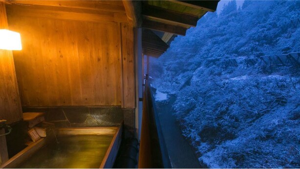 A wooden interior with a wooden bench and a small table, a wooden wall with a light fixture, and a view of a snow-covered landscape outside. - Sake Hotel Tamakiya (Tokamachi)