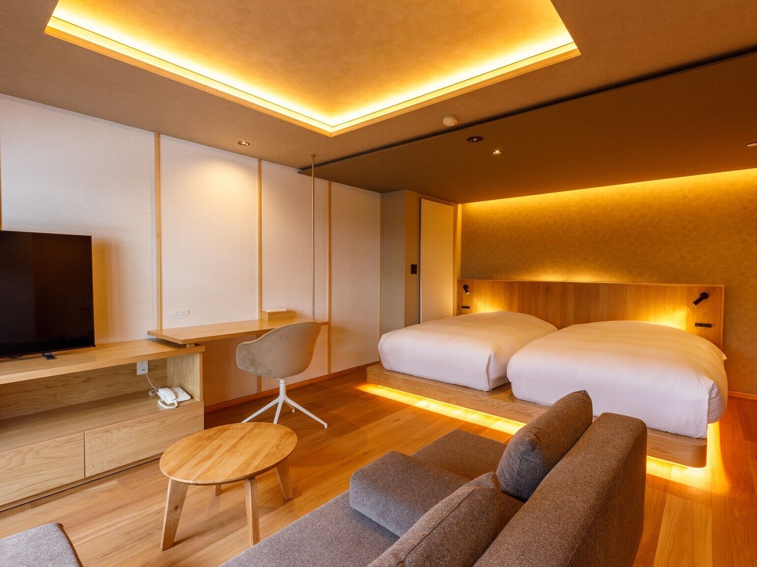 A modern hotel room with a flat-screen TV, a desk, a chair, a sofa, and two beds.
