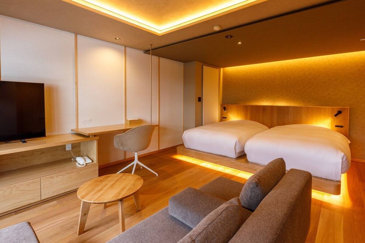 A modern hotel room with a flat-screen TV, a desk, a chair, a sofa, and two beds.