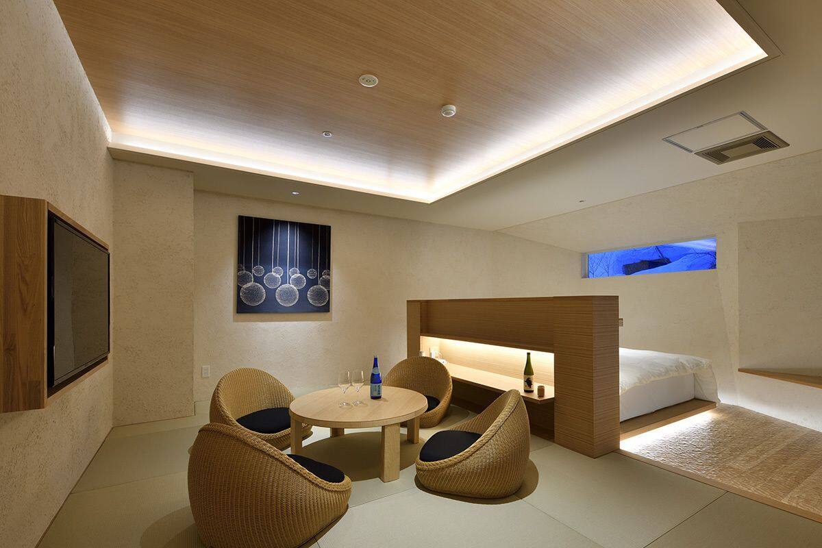 A modern hotel room with a round dining table, four wicker chairs, a sofa, a fireplace, and a large TV screen.