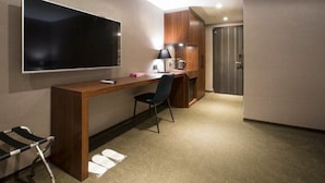 Et hotelværelse med et stort fladskærms-tv på væggen, et træskrivebord med stol, et lille bord med lampe og et garderobeskab.