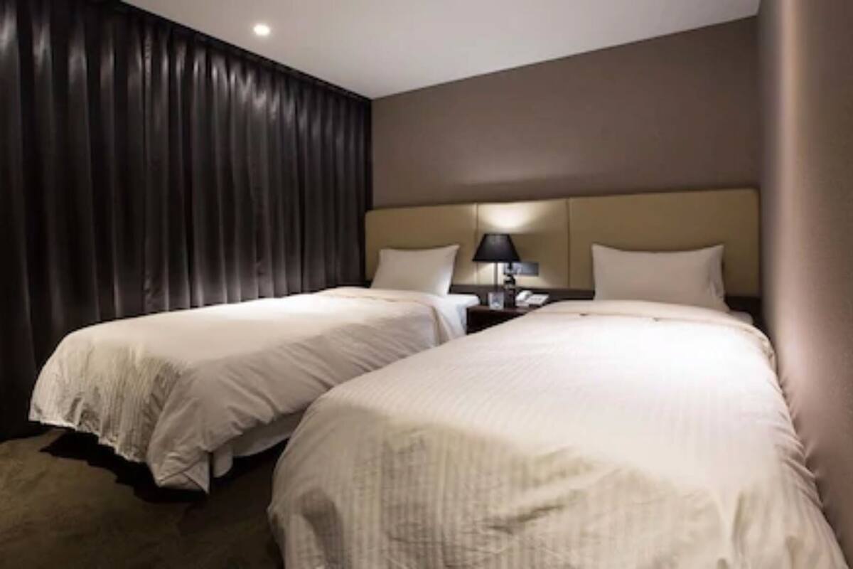 A hotel room with two beds, dark curtains, a nightstand with a lamp, and a neutral color scheme.