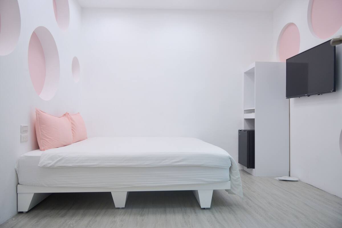 A modern bedroom with a white bed, pink pillows, a flat-screen TV mounted on the wall, and a minimalist design.
