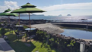 An outdoor seating area with tables and chairs, green umbrellas, and a pool with a curved edge.