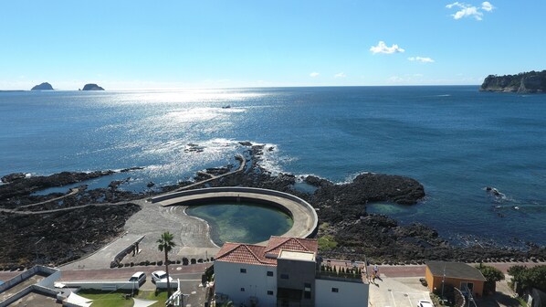 A coastal area with a circular pool, rocky shoreline, and buildings with red-tiled roofs.