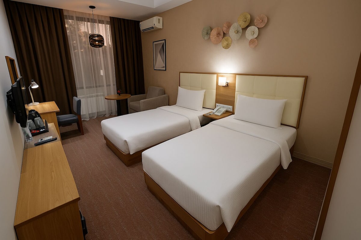 A hotel room with two single beds, a desk with a chair, a small table, a lamp, and a wall-mounted phone.