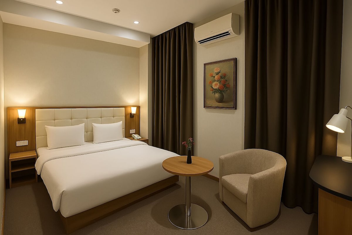 A hotel room with a large bed, bedside tables, a round table, a chair, an air conditioning unit, and a painting on the wall.