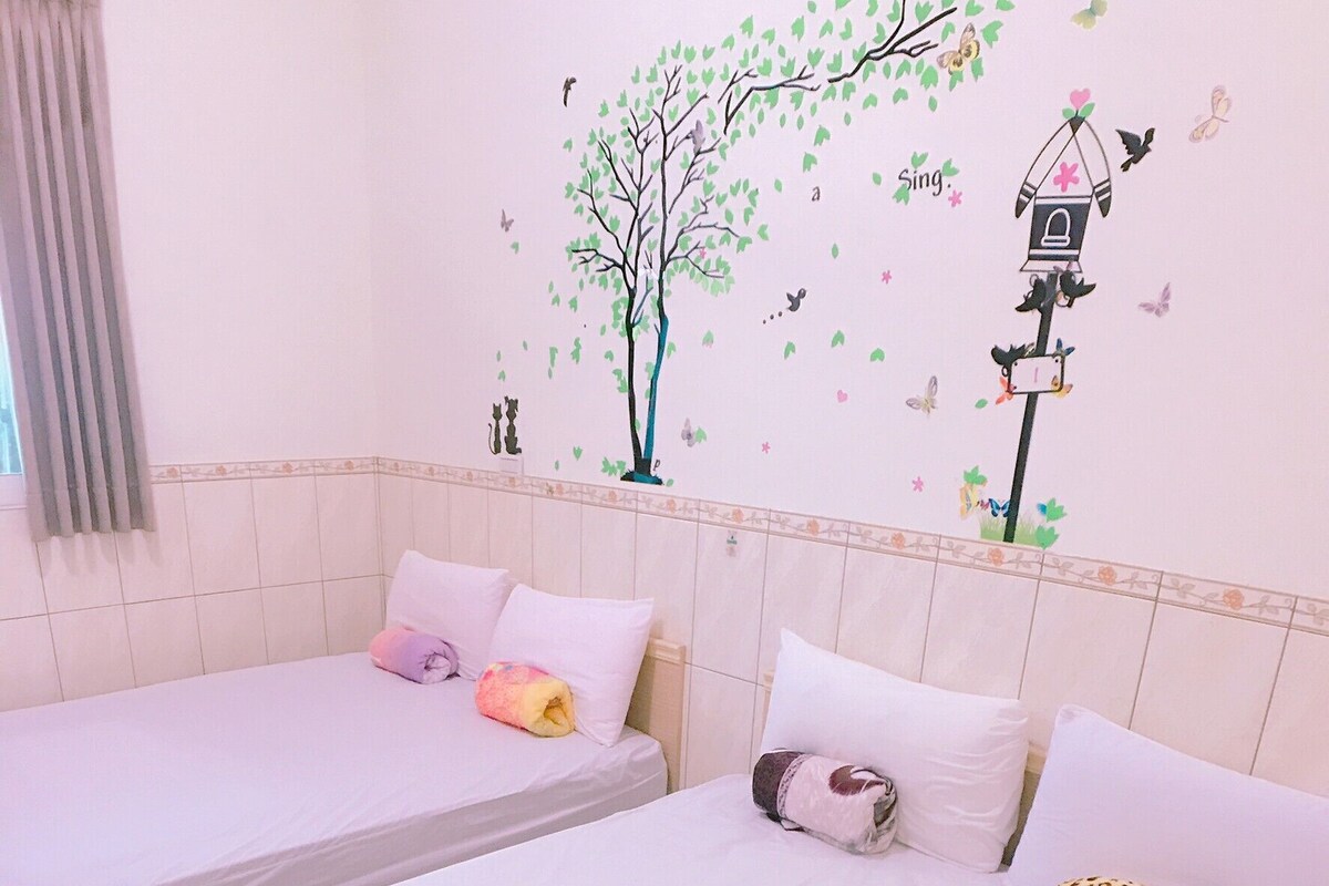 A child lying on a bed with a checkered bedspread. The room has two beds with white bedding and decorative pillows. There is a wall decal of a tree and birds.