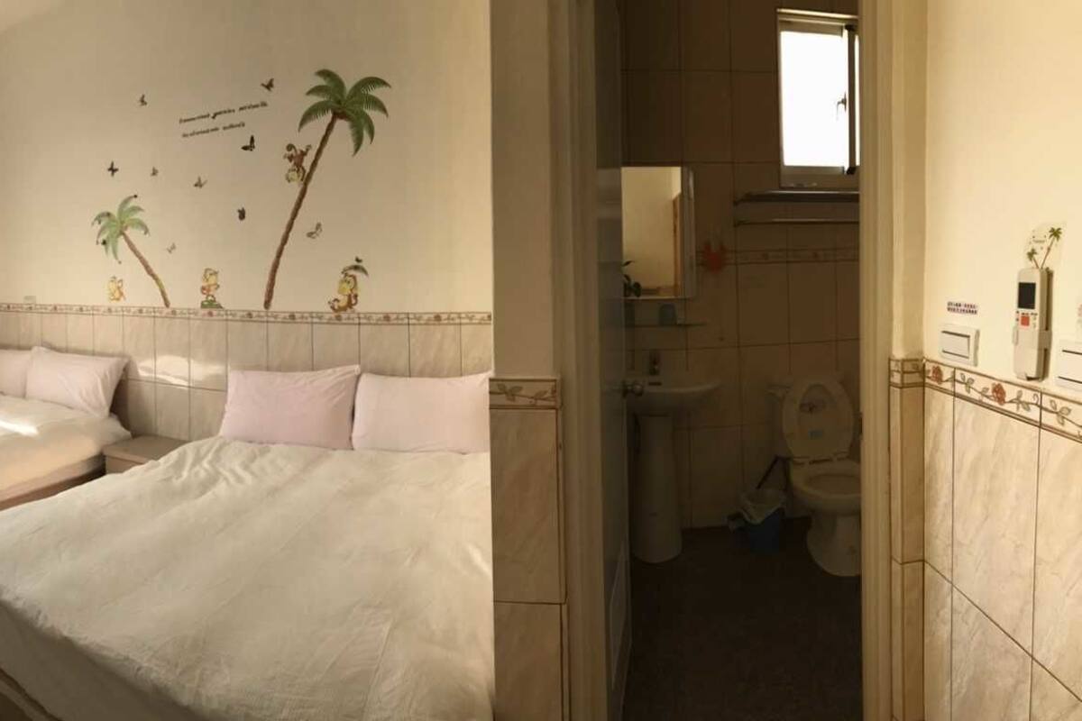 A bedroom with two beds, a window with curtains, a wall-mounted air conditioner, and a bathroom visible through an open door.