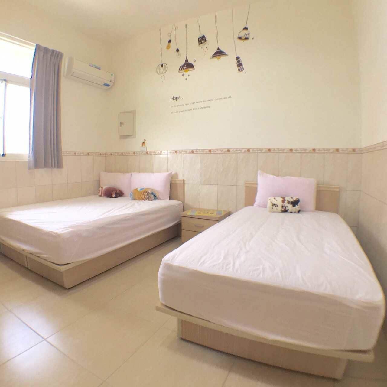 Two single beds with white linens, a small bedside table with a book, and a wall-mounted air conditioner.