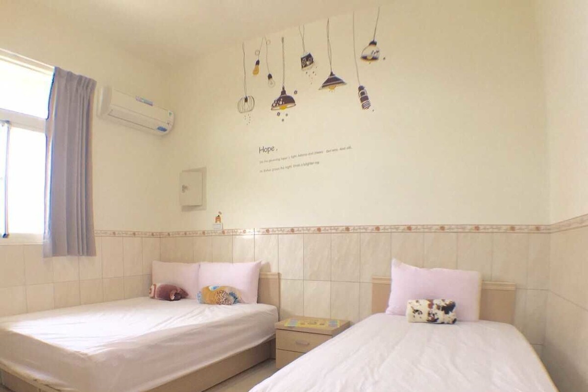 Two single beds with white linens, a small bedside table with a book, and a wall-mounted air conditioner.