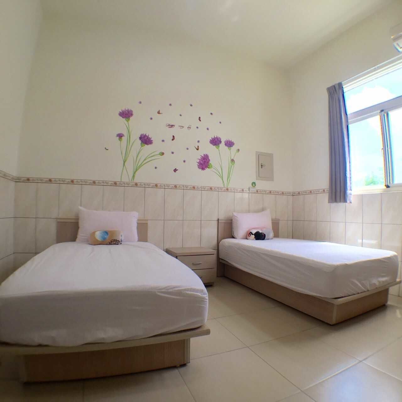 Habitación de hotel con dos camas, una ventana con cortinas y un adorno de flores moradas en la pared.