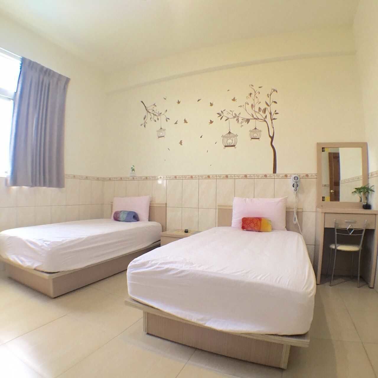 A hotel room with two single beds, a desk with a chair, a mirror, and a wall decoration of birds and a tree.