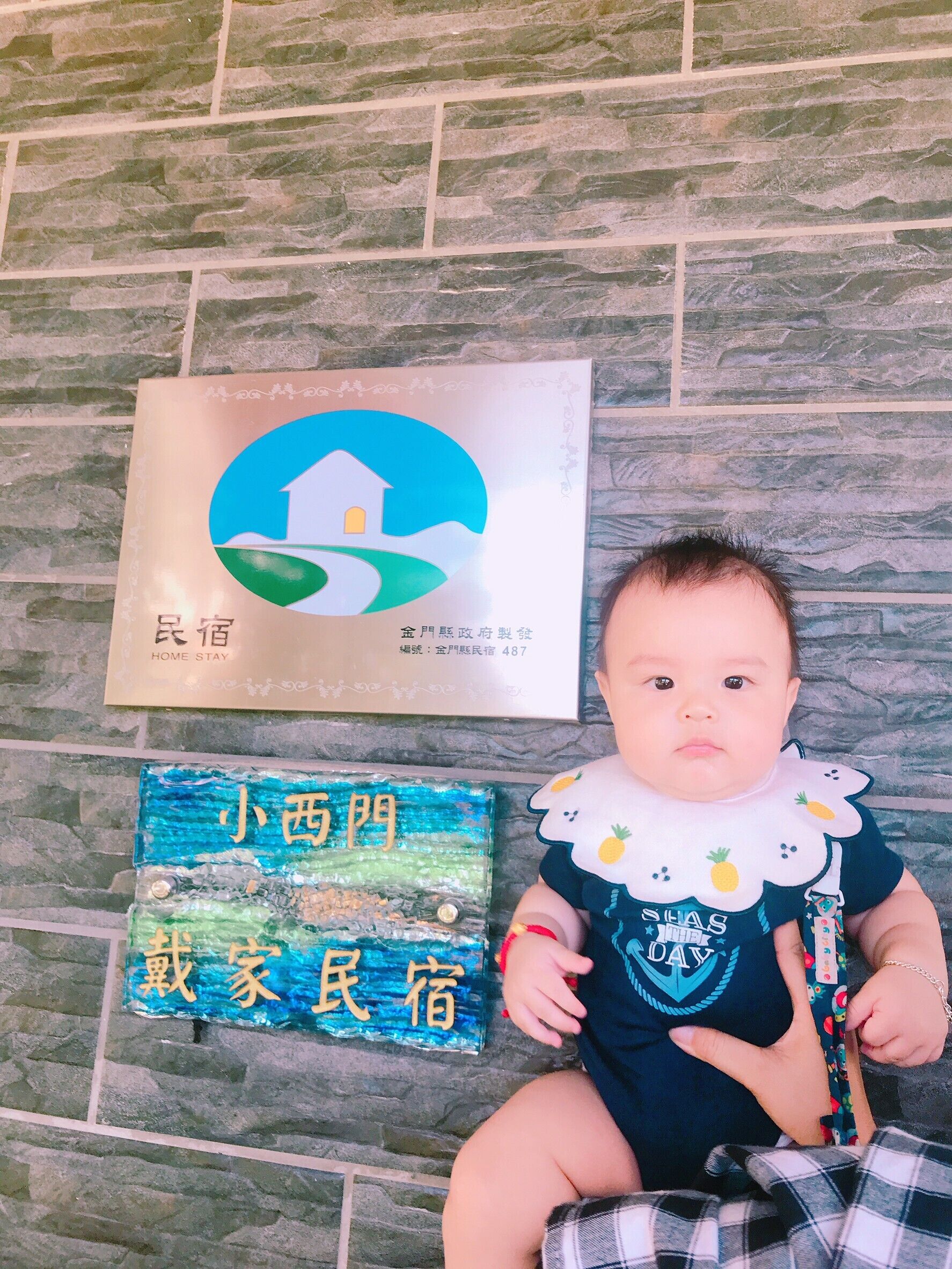 A baby sitting on a stone floor with a placard displaying a house logo and text, and another placard with text in a foreign language.