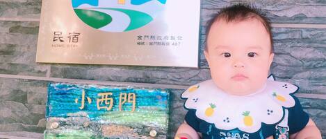 A baby sitting on a stone floor with a placard displaying a house logo and text, and another placard with text in a foreign language.
