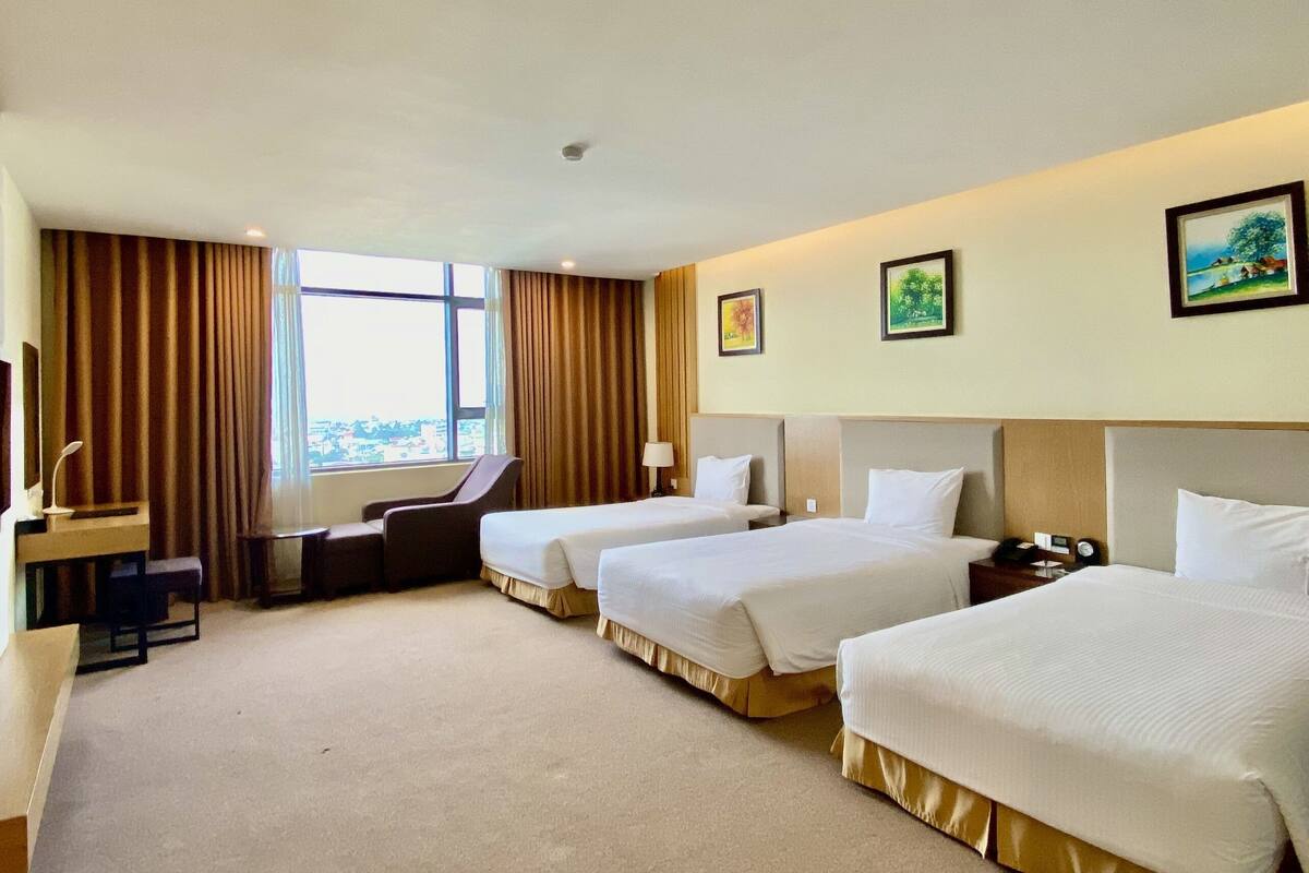 A hotel room with three single beds, a desk with a chair, a large window with curtains, and two framed pictures on the wall.