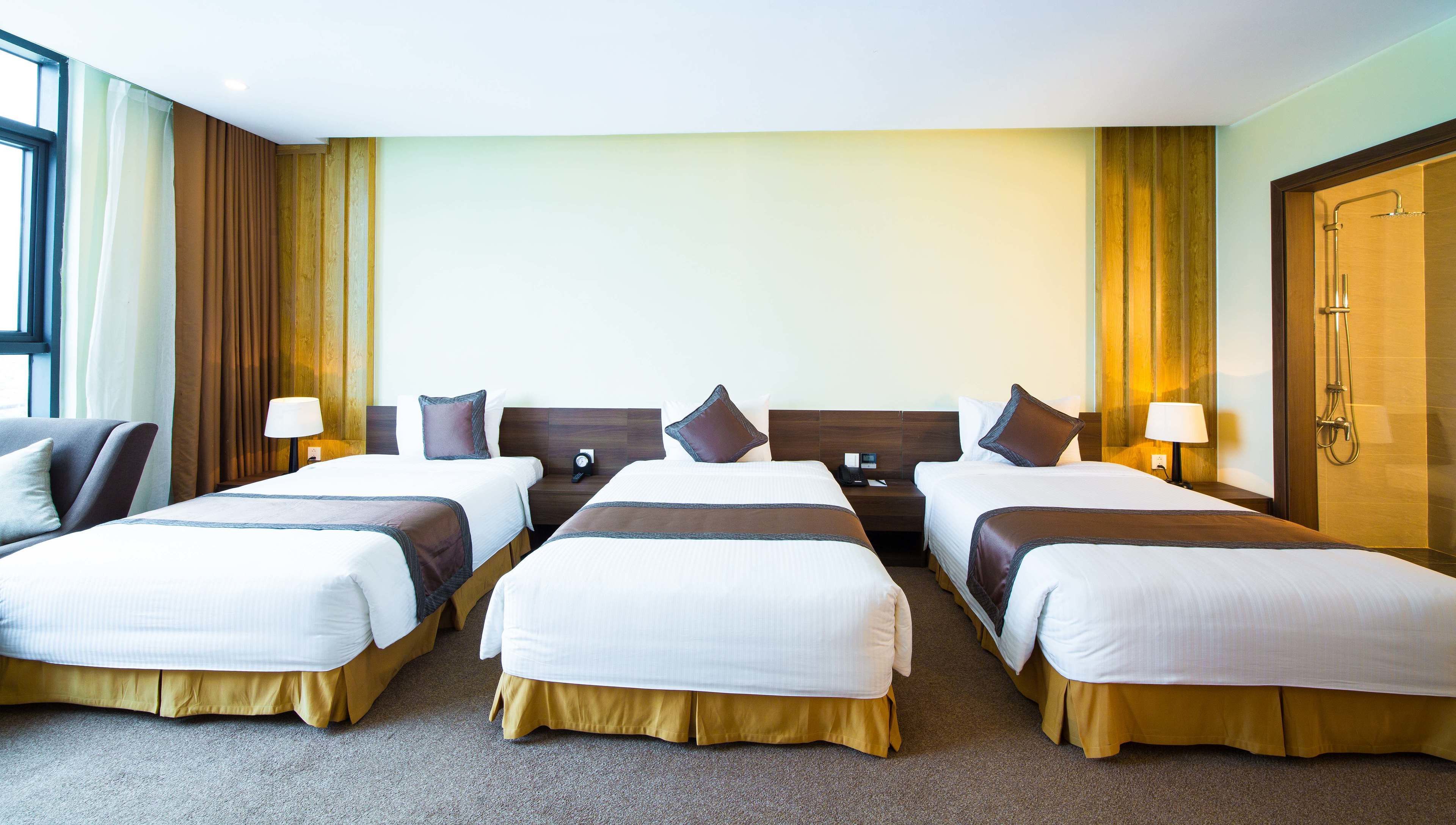 Habitación de hotel con tres camas, cada una con un corredor marrón, ropa de cama blanca y cabeceros de madera. Se ve una puerta de madera a la derecha y una ventana a la izquierda.