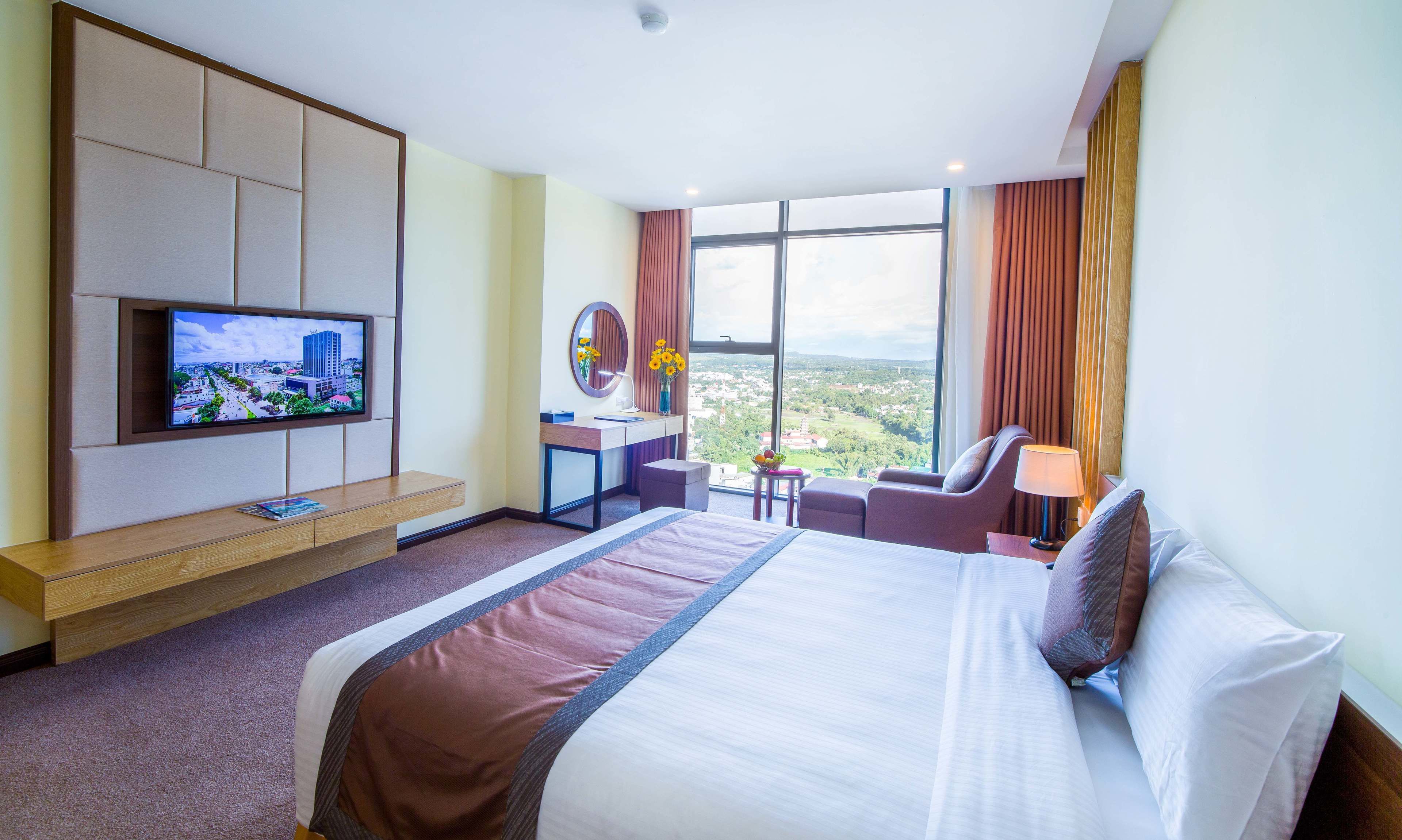A hotel room with a large bed, a desk with a computer, a TV mounted on the wall, and a large window offering a city view.