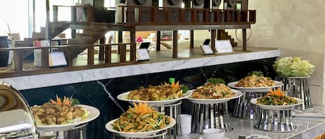Un área de bufé con varias estaciones de comida, un mostrador de recepción y una estructura con techo de paja.