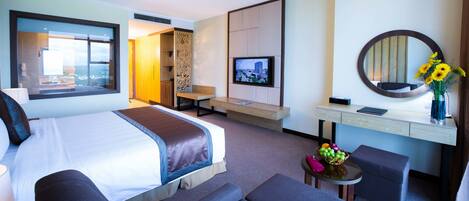 A hotel room with a large bed, a desk with a computer, a TV, a mirror, and a vase with flowers.