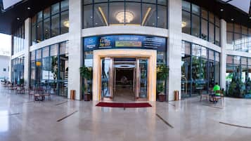 A modern hotel lobby with a glass entrance, seating areas, and a red carpet leading to the reception.