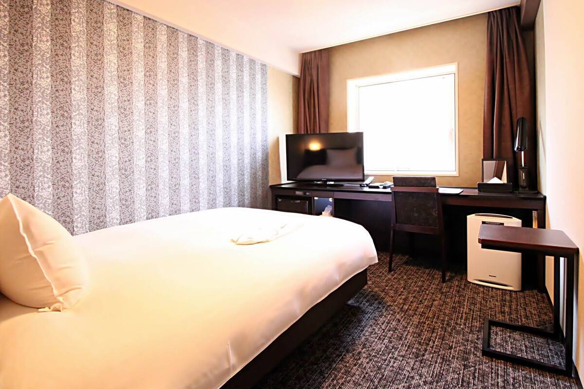 Ein Hotelzimmer mit einem großen Bett, einem Schreibtisch mit Fernseher und einem Fenster mit Vorhängen.