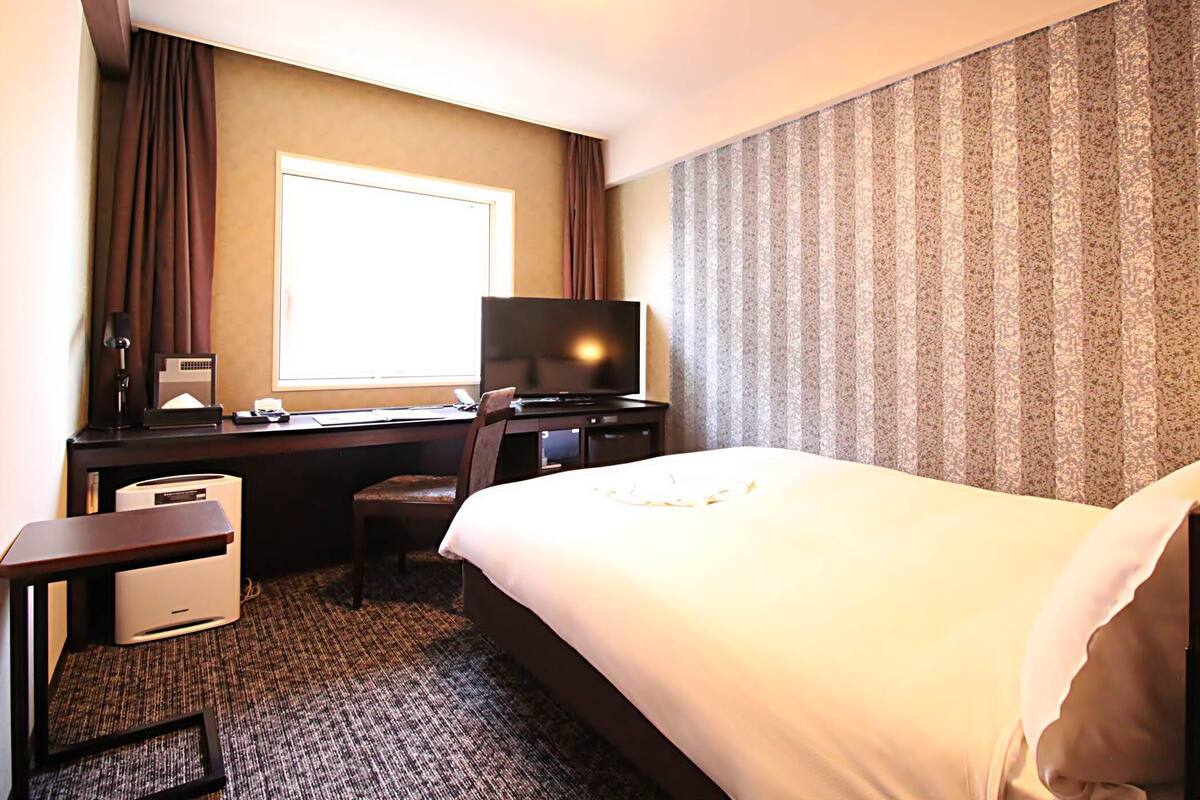 Ein Hotelzimmer mit Bett, Schreibtisch, Computer, Fernseher und einem Fenster mit Vorhängen.