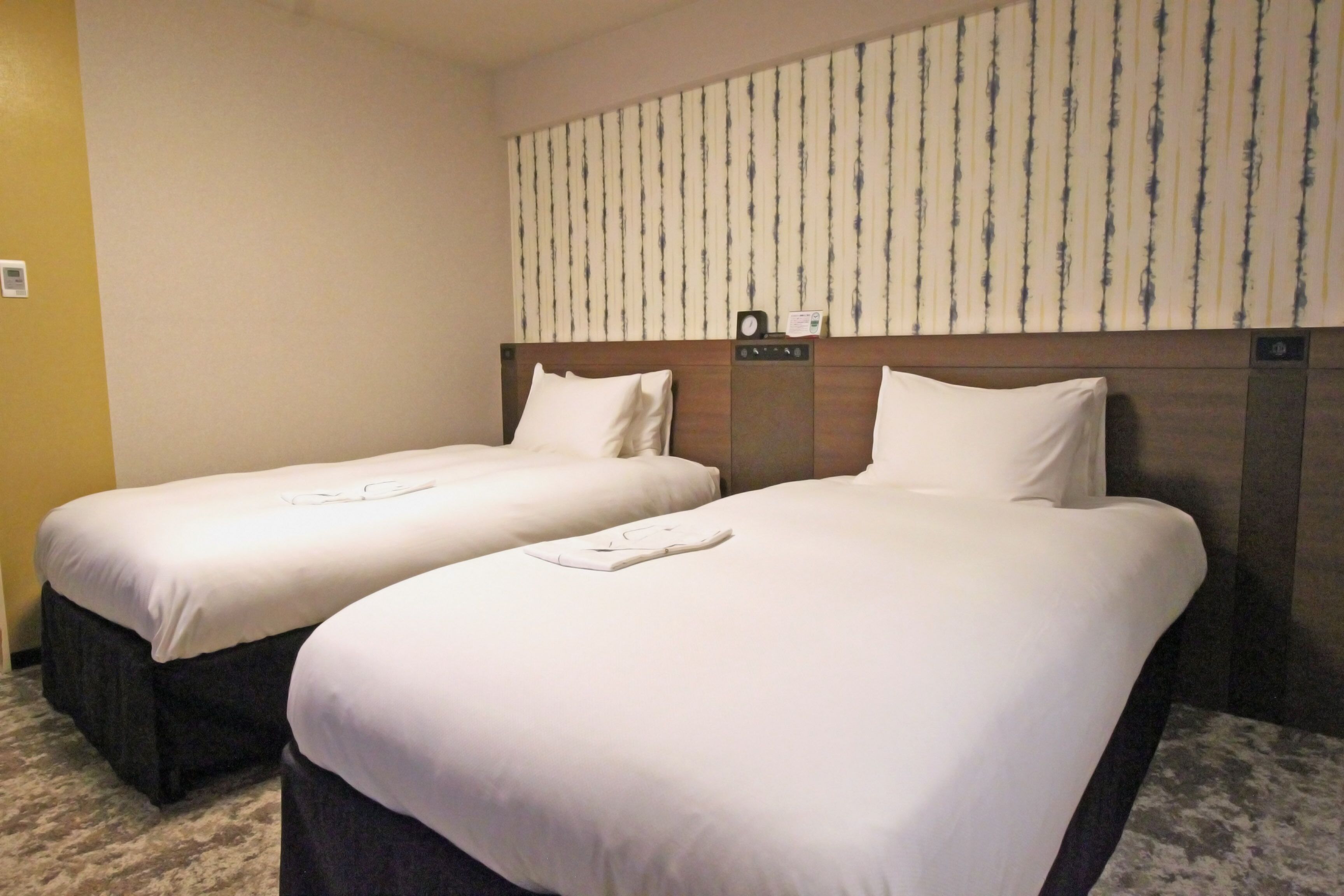 A hotel room with two single beds, a headboard with a clock, and a striped wallpaper on the wall.