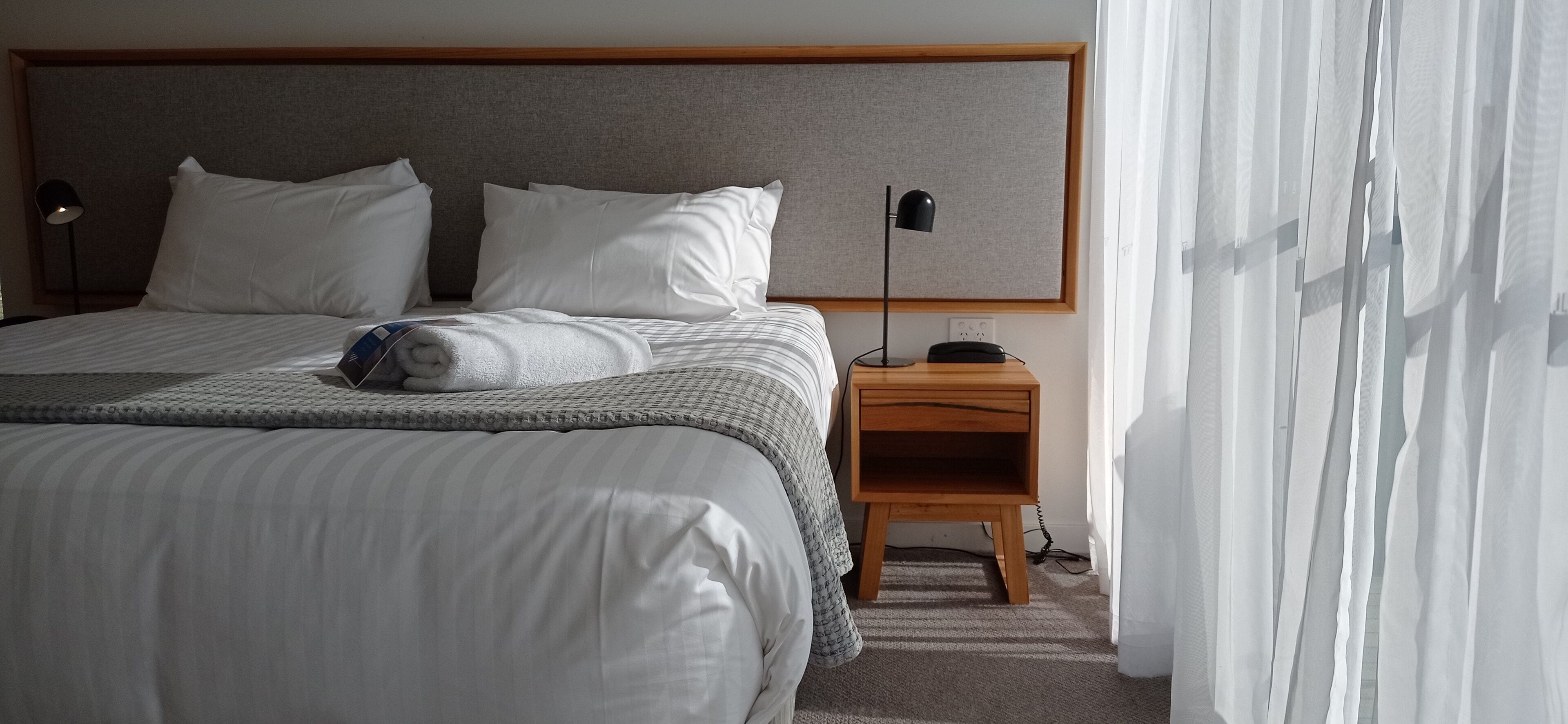 A neatly made bed with white linens, a bedside table with a lamp, and a power outlet on the wall.