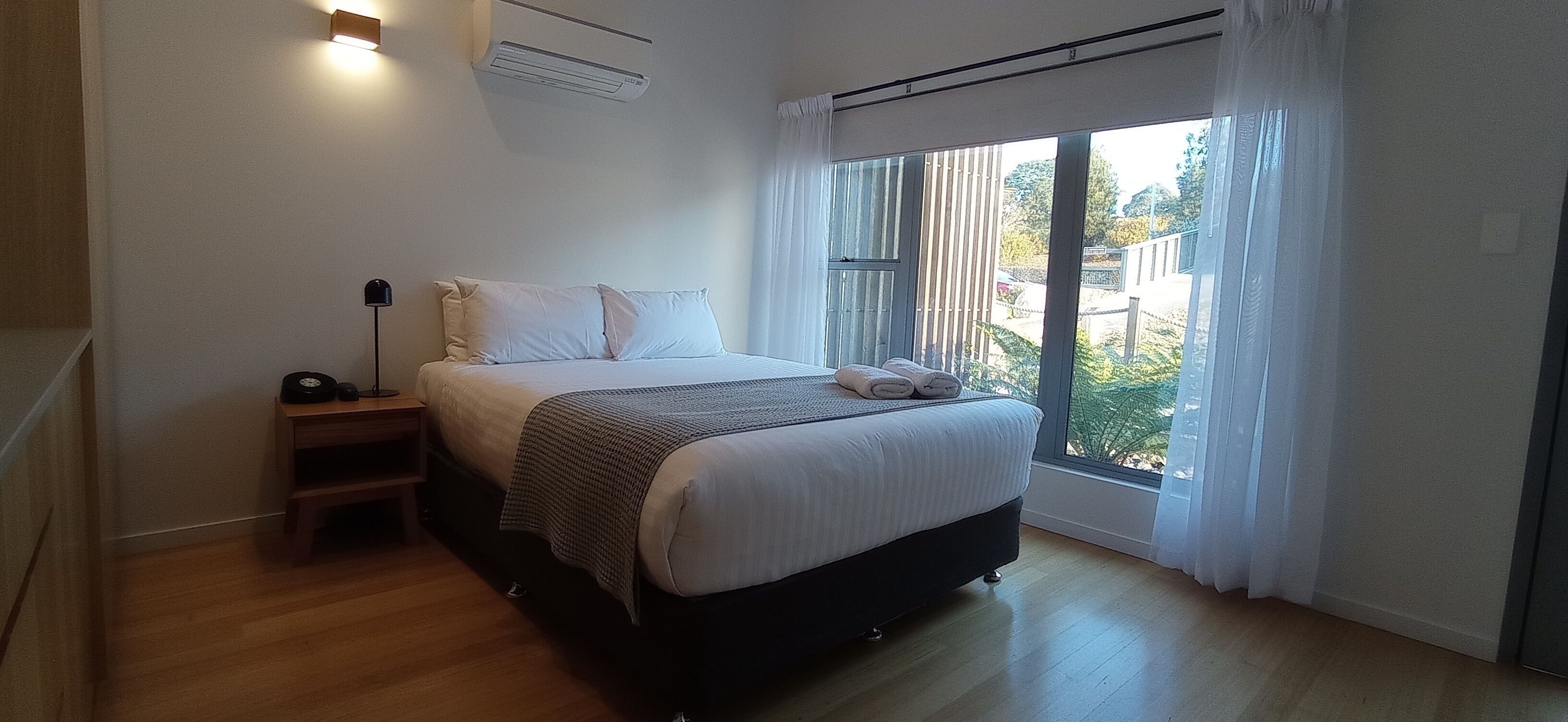 A bedroom with a bed, bedside table, lamp, and a view of the outdoors through a large window.