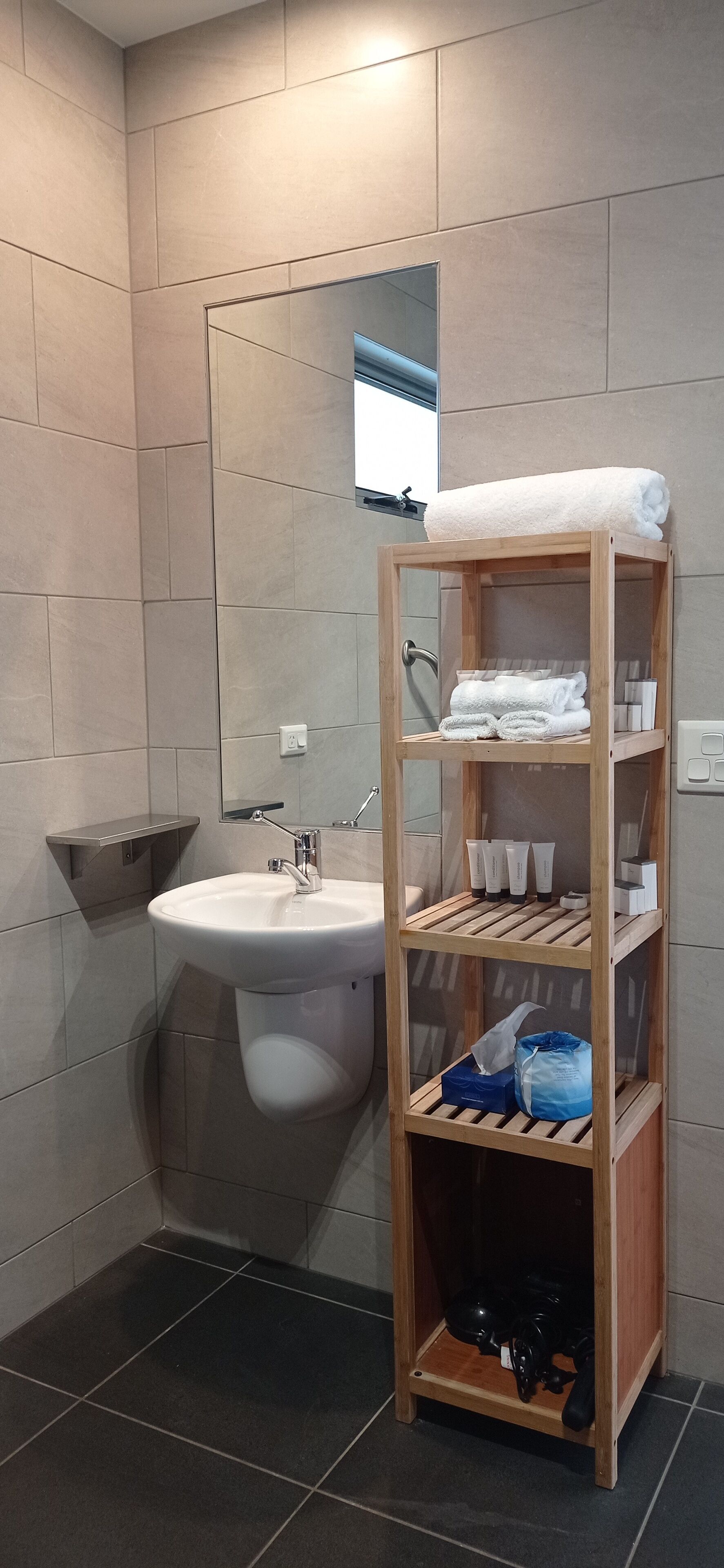 Une salle de bain moderne avec une étagère en bois où sont rangées des serviettes, des articles de toilette et un sac bleu.