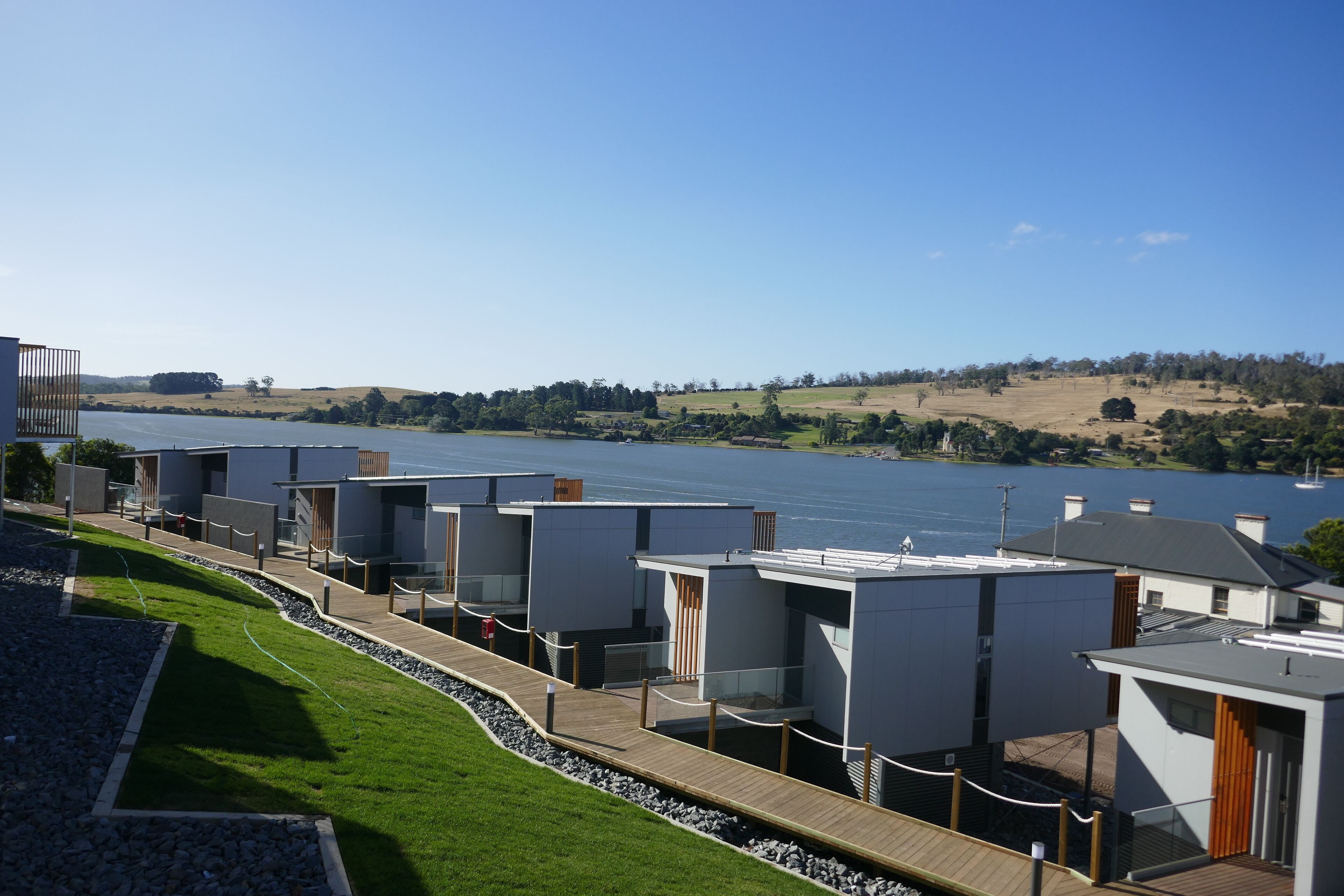 Una hilera de cabañas modernas y compactas, con terraza de madera que da a un cuerpo de agua, rodeadas de zonas ajardinadas.