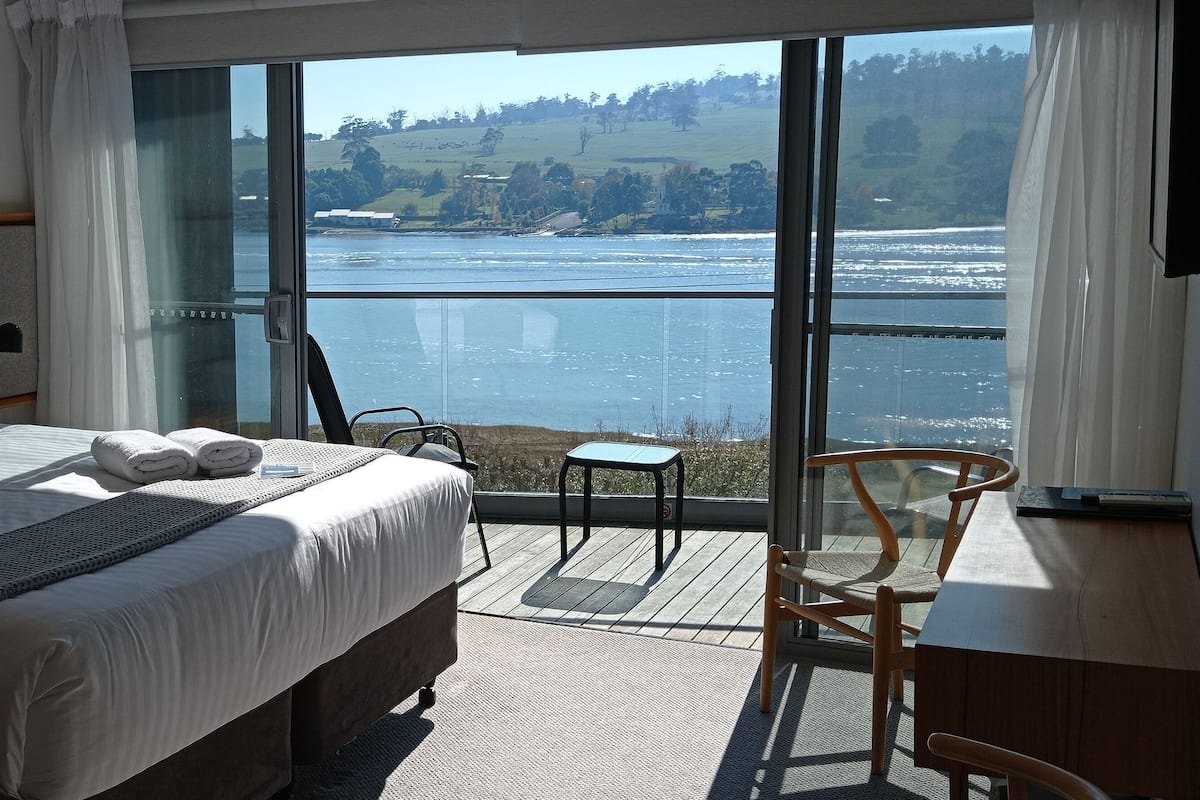 Ein Hotelzimmer mit einem großen Bett, einem kleinen Tisch, einem Sessel und Blick auf einen See.
