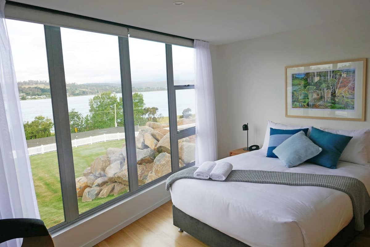 Ein Schlafzimmer mit großem Fenster, einem Bett mit Kissen, einem Bild an der Wand und Blick auf einen See und Felsen.