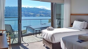 Een hotelkamer met twee bedden, een balkon met uitzicht op een meer en heuvels, en een tafel met stoelen.