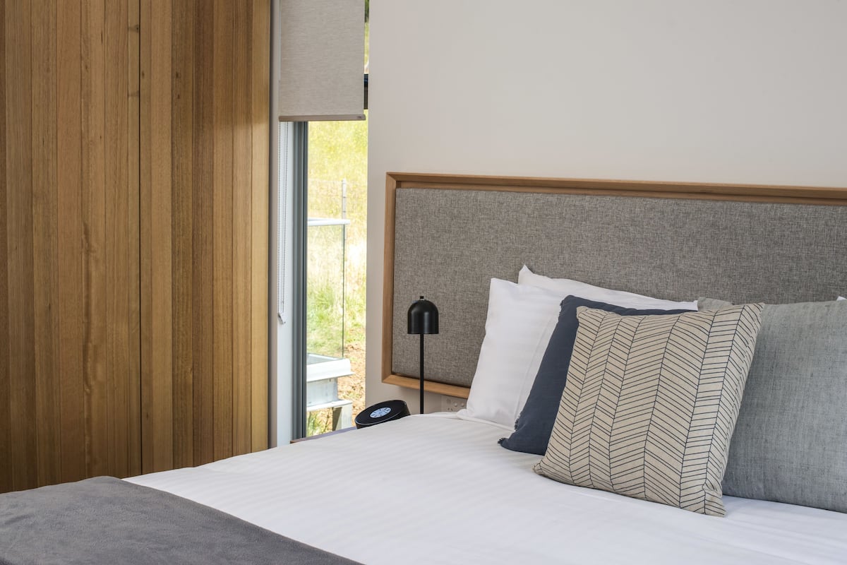 Ein Schlafzimmer mit Holzschrank, einem Bett mit vielen Kissen, einem Nachttisch mit Lampe und einem Fenster mit Blick ins Grüne.