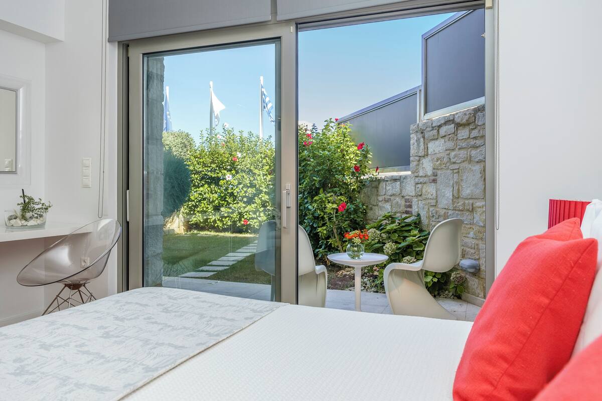 A modern room with a glass door leading to a garden, a white bed, a chair, and a small table with a vase.