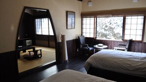 A hotel room with two beds, a small table, a chair, a desk, and a large window with a view of trees.