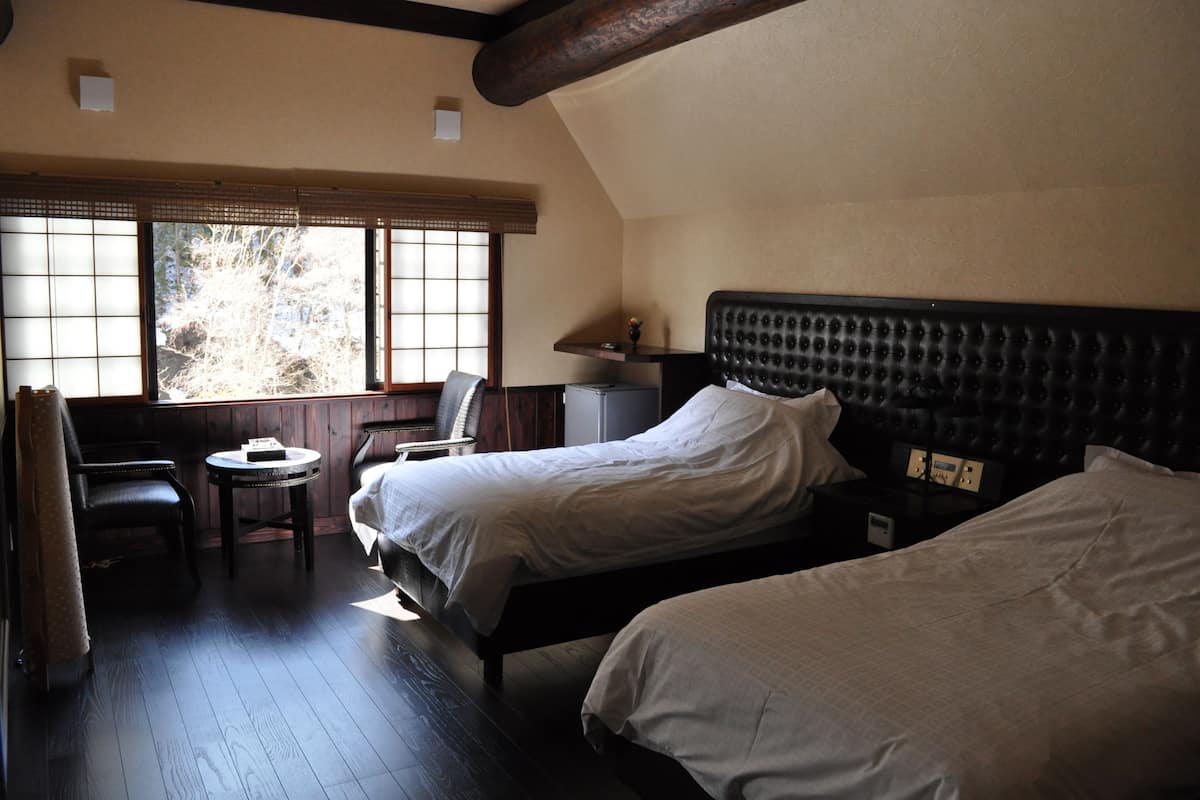 A room with two beds, a small table, a chair, and a window with a view of trees.