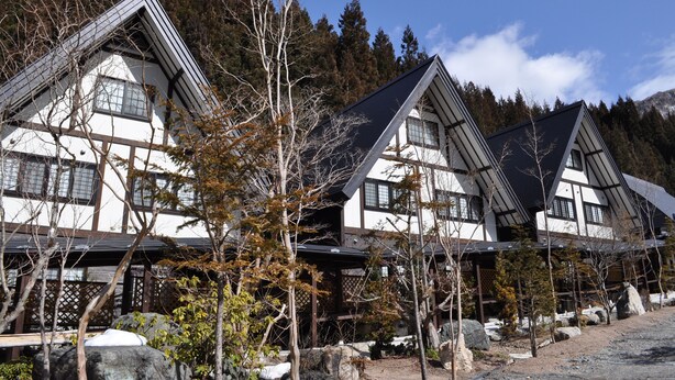 A row of traditional Japanese-style buildings with steeply pitched roofs, surrounded by trees and mountains.
