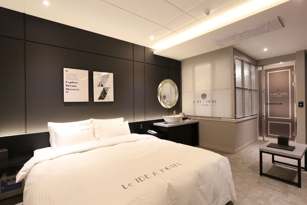 Habitación de hotel con una cama grande, una mesita de noche con lámpara, un espejo y dos citas enmarcadas en la pared.