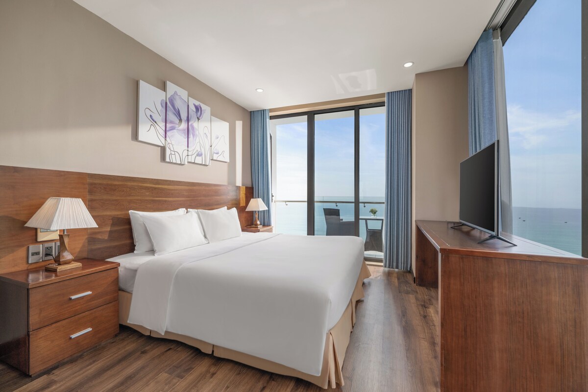 A hotel room with a large bed, a TV, a desk, and a balcony with a view of the sea.