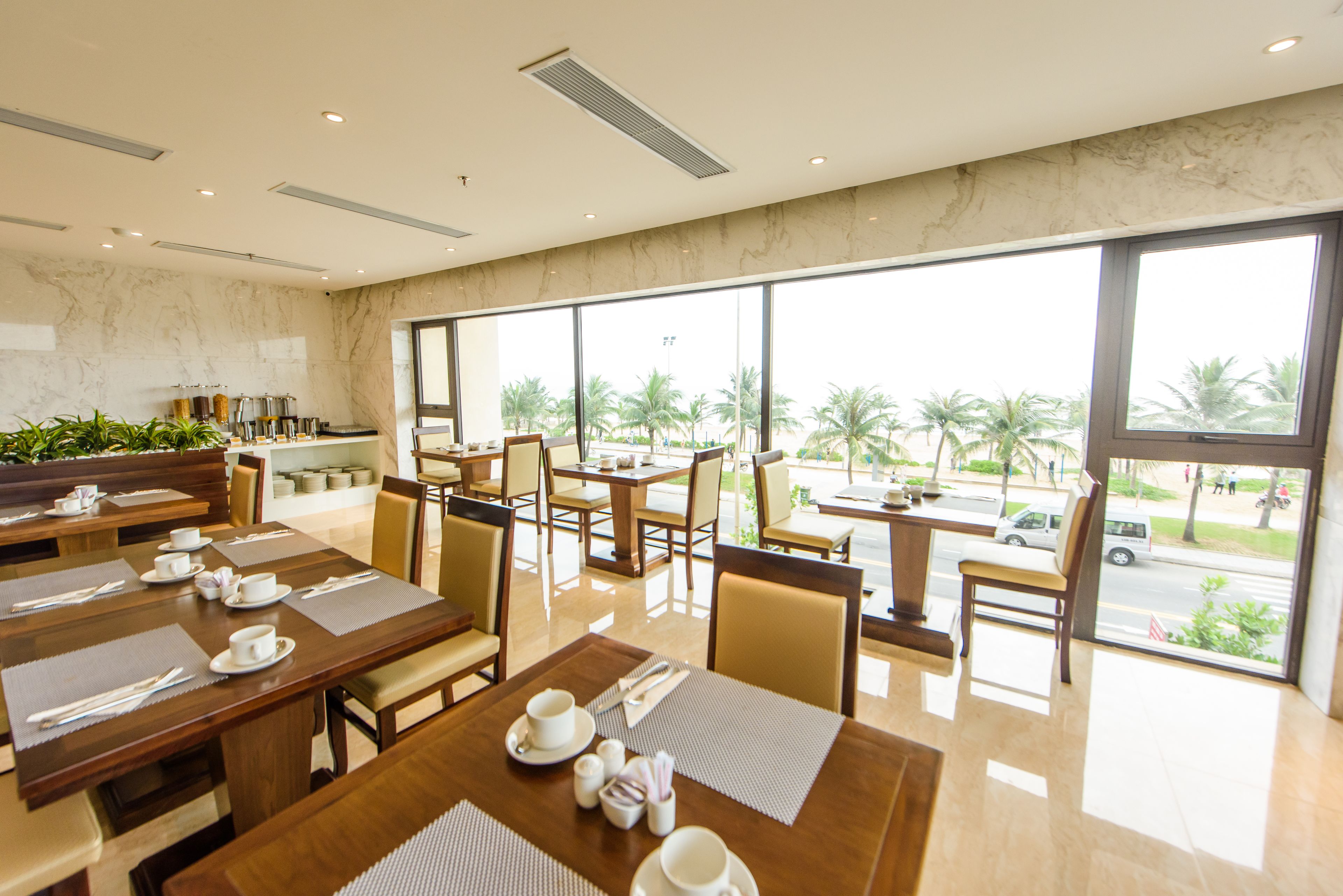 Une salle à manger d’hôtel avec de nombreuses tables préparées pour le petit-déjeuner, de grandes fenêtres offrant une vue sur des palmiers et une route, et un mur en marbre.